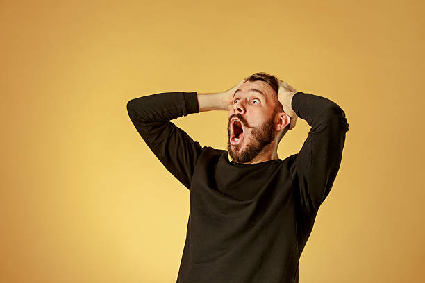 Studio shot of a bearded man against an ochre backdrop, hands at his head and mouth wide open.